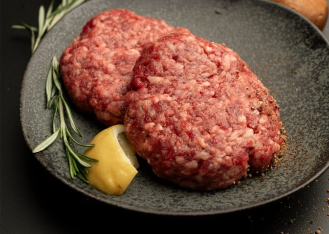Fire Up the Grill! 5 Best-Quality Meat Delivery Services in the South - 2 Two raw ground beef patties on a dark plate with a sprig of rosemary, a dollop of mustard, and a lemon wedgeβperfect for the grill or as inspiration for your next order from meat delivery services in the South.