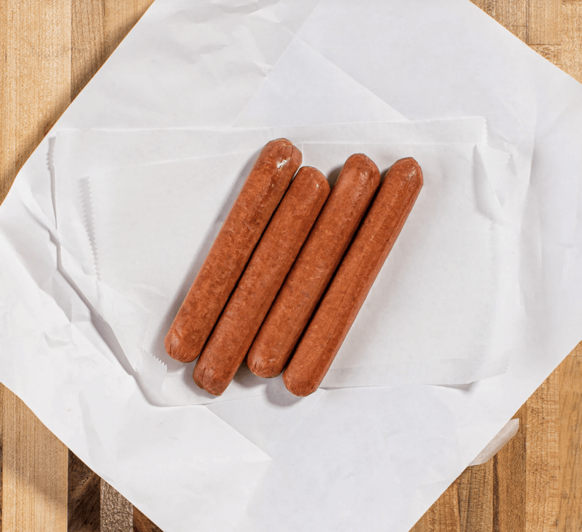 Fire Up the Grill! 5 Best-Quality Meat Delivery Services in the South - 4 Four uncooked hot dogs, perfect for the grill, are arranged side by side on a sheet of white parchment paper atop a wooden surface.