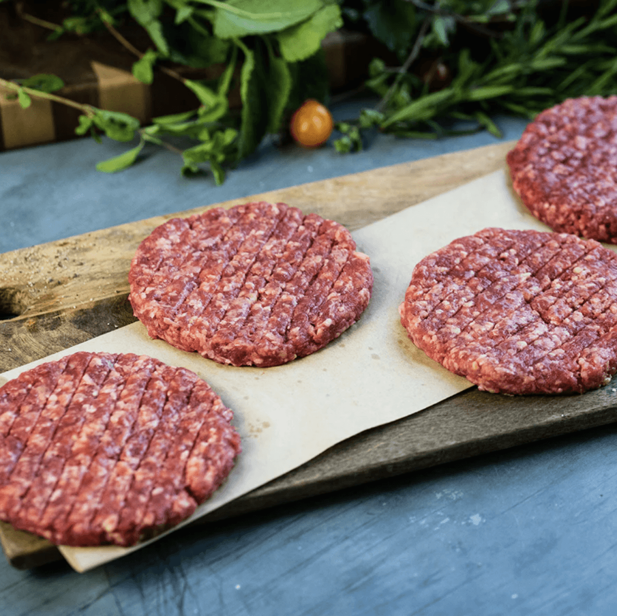 Fire Up the Grill! 5 Best-Quality Meat Delivery Services in the South - 5 Four raw beef burger patties on parchment paper atop a wooden board, ready for the grill, with fresh herbs and vegetables in the backgroundβa classic taste of the South now made even easier with premium meat delivery services.