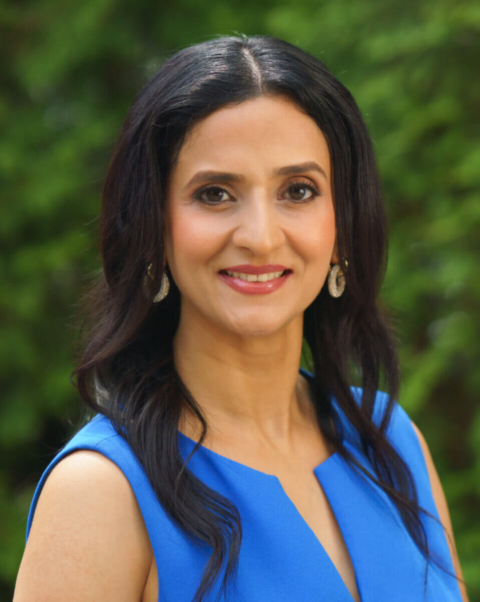 This Nashville Doctor Helps Families Disconnect to Reconnect - 2 A woman with long dark hair, wearing a sleeveless blue dress and earrings, smiles at the camera in an outdoor setting with a green blurred background.