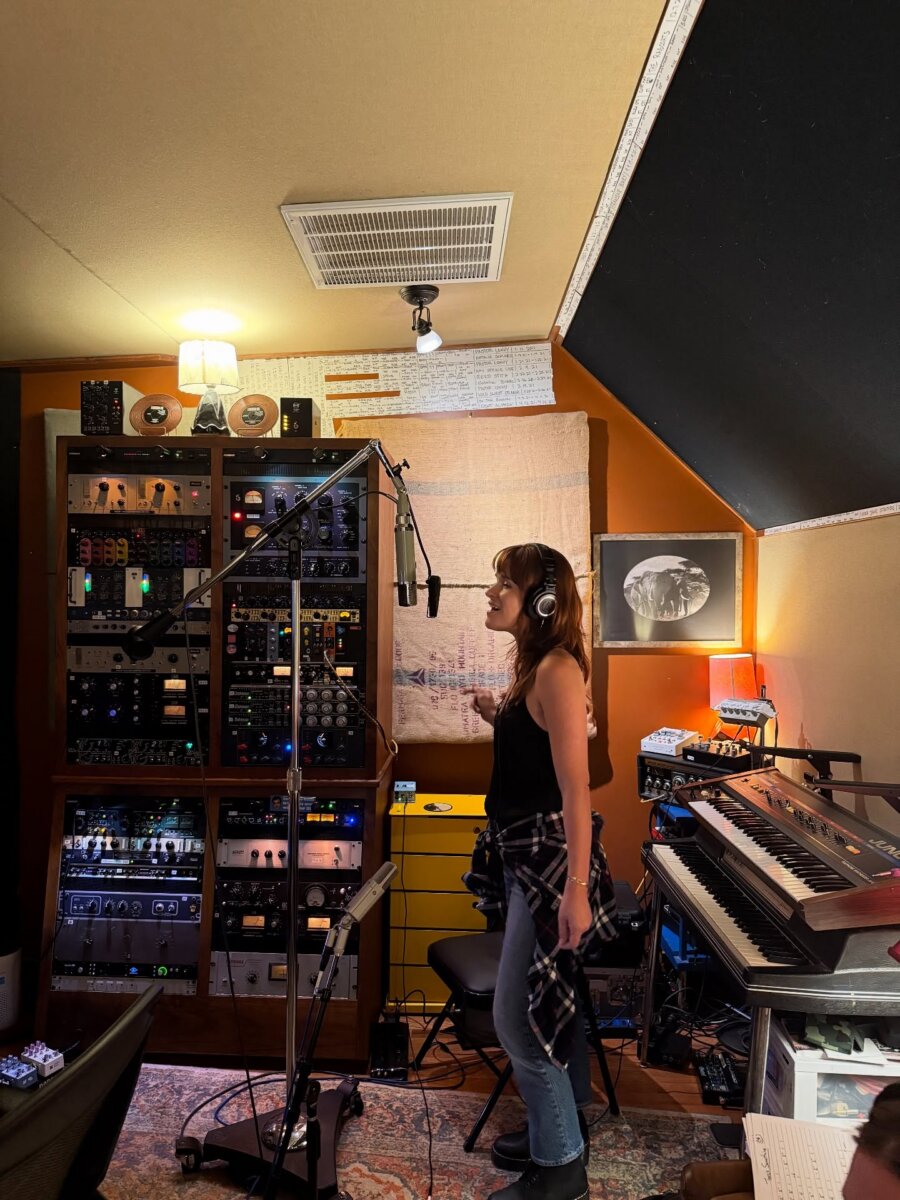 Singer-Songwriter Lacy Green is Turning Life into Lyrics - 4 A woman wearing headphones sings into a microphone in a cozy music studio filled with audio equipment, a keyboard, and warm lighting, reminiscent of Lacy Greenβs intimate recording sessions.