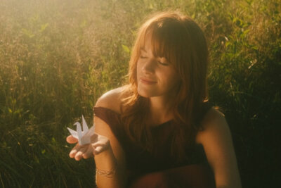 A woman with red hair sits in a sunlit, grassy field in Lacy Green, smiling softly and holding a white paper crane in her hand.