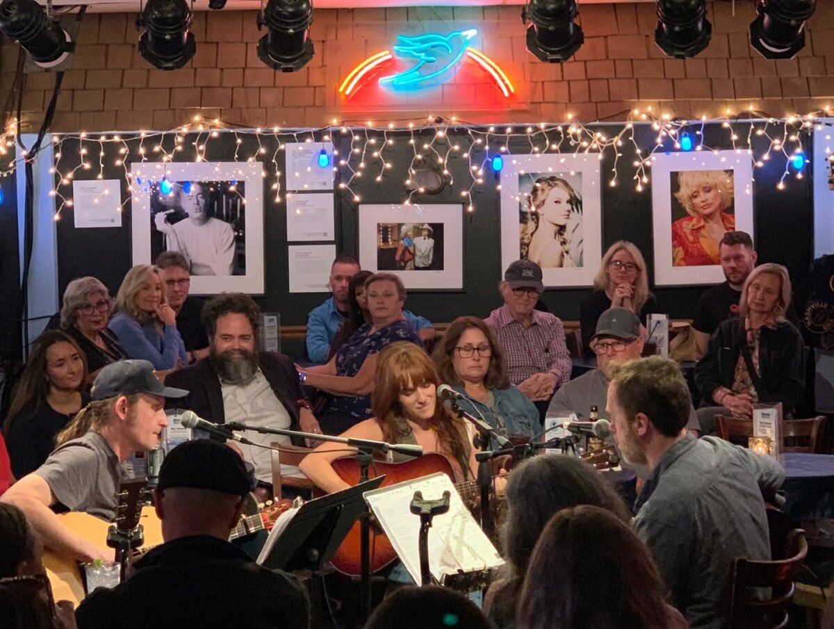 Singer-Songwriter Lacy Green is Turning Life into Lyrics - 6 Musicians, including Lacy Green, perform with guitars for an attentive audience in a cozy, art-filled venue with string lights and a neon bird sign overhead.