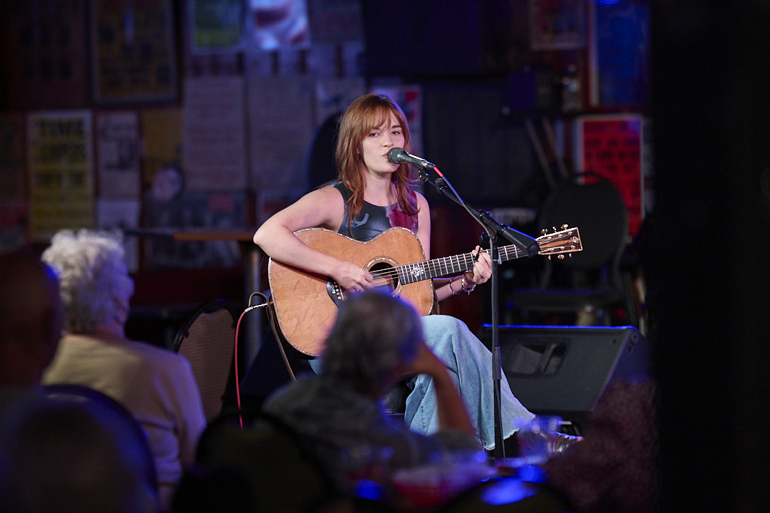 Singer-Songwriter Lacy Green is Turning Life into Lyrics - 2 Lacy Green sits on stage playing an acoustic guitar and singing into a microphone, with an audience seated and watching her performance in a dimly lit venue.