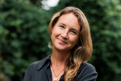 Varina Buntin Willse, with long, light brown hair, smiles softly while standing outdoors in front of blurred green foliage, wearing a dark shirt and layered necklaces.