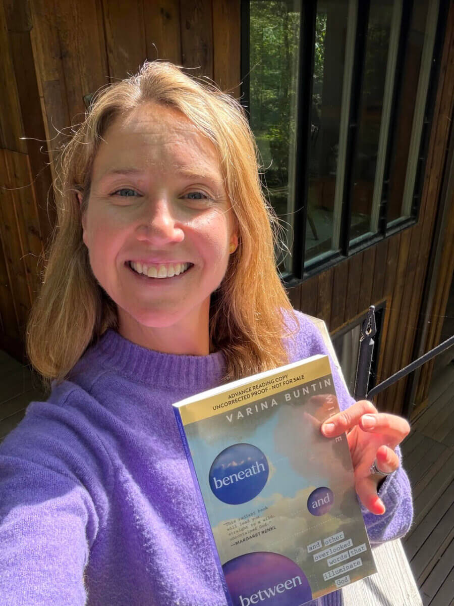 This Nashville Author Sees the Extraordinary in Ordinary Words - 3 A woman in a purple sweater smiles and holds a copy of "beneath and between" by Varina Buntin Willse outside a wooden building.