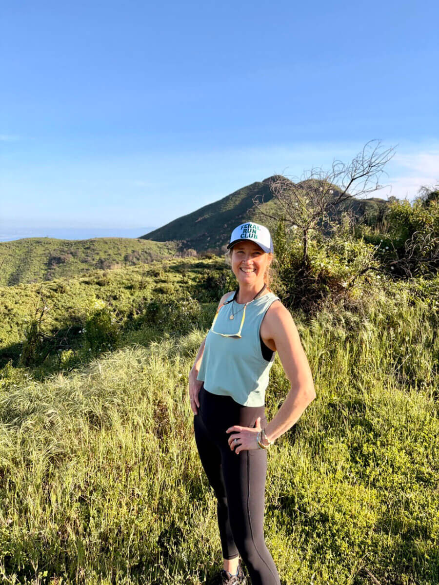 This Nashville Author Sees the Extraordinary in Ordinary Words - 5 Varina Buntin Willse stands smiling on a grassy hillside, wearing a light blue tank top, black leggings, and a cap, with rolling green hills and a blue sky in the background.