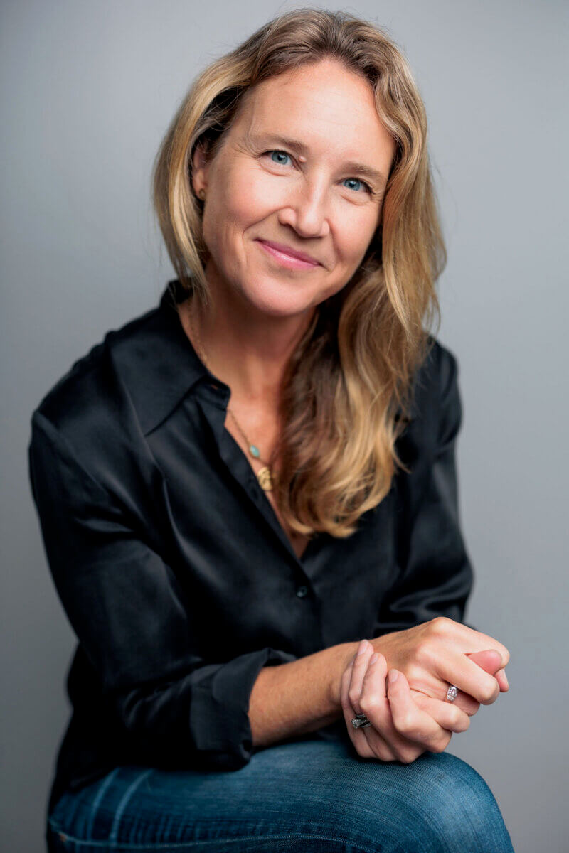 This Nashville Author Sees the Extraordinary in Ordinary Words - 2 Varina Buntin Willse, a woman with long blonde hair wearing a black shirt and blue jeans, sits and smiles at the camera against a plain grey background.