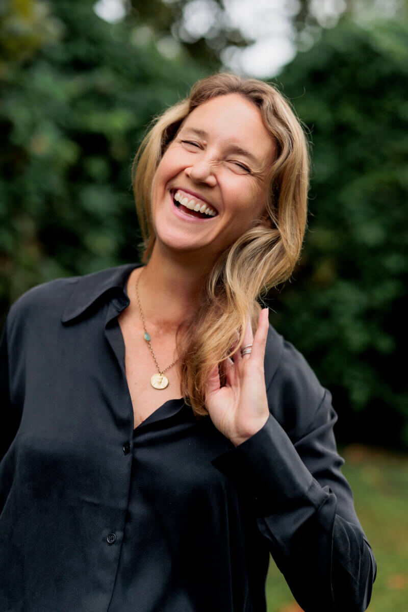 This Nashville Author Sees the Extraordinary in Ordinary Words - 7 Varina Buntin Willse, a woman with long blonde hair wearing a black button-up shirt and gold necklace, smiles and holds her hand up while standing outdoors with greenery in the background.