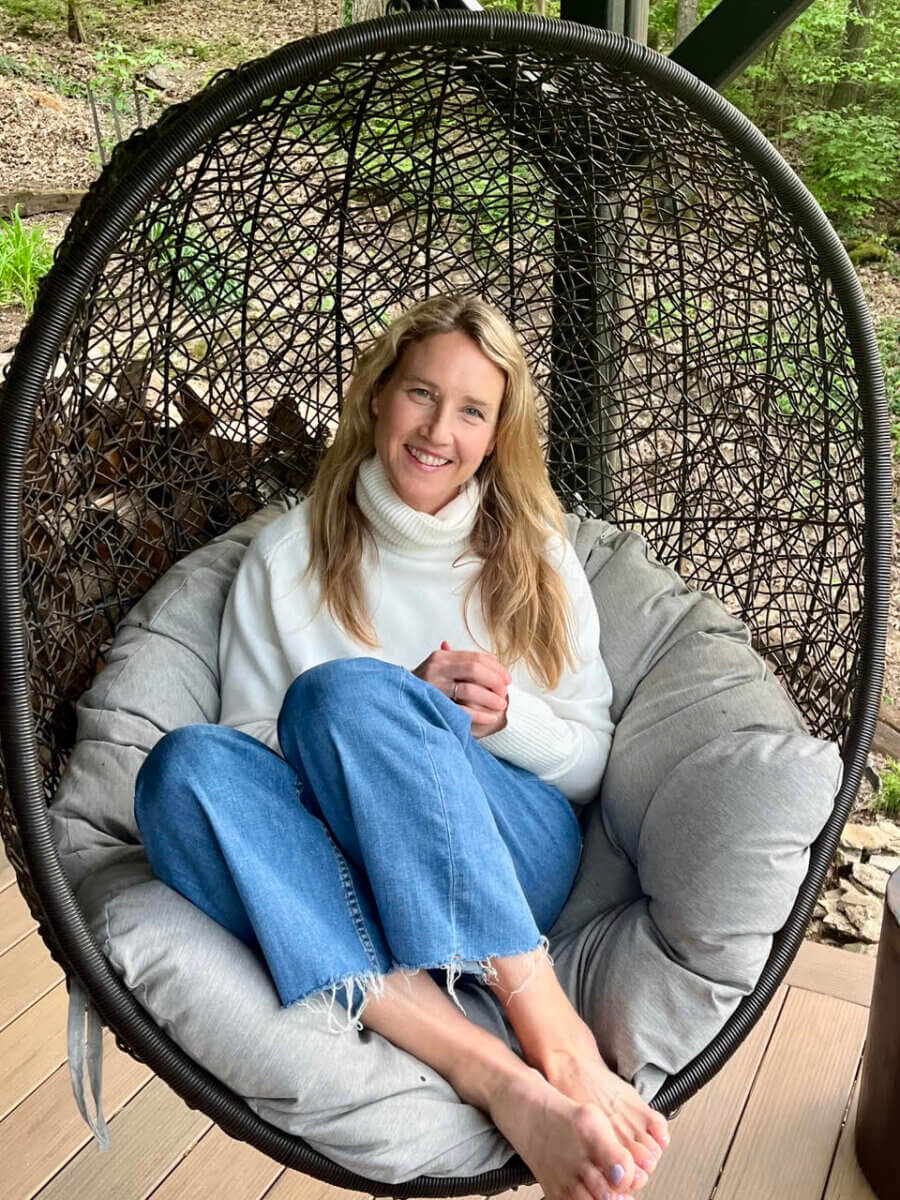 This Nashville Author Sees the Extraordinary in Ordinary Words - 6 Varina Buntin Willse, wearing a white sweater and blue jeans, sits barefoot in a hanging egg chair outdoors, smiling at the camera.