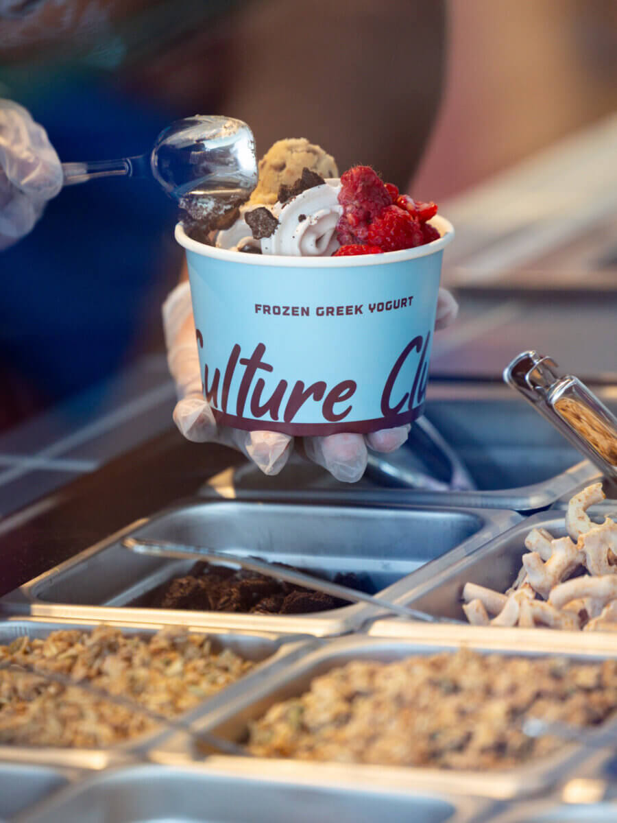 A person scoops toppings onto a cup of frozen Greek yogurt at a self-serve counter—one of the popular features in new restaurants opening around Nashville. An array of toppings is visible in metal containers.