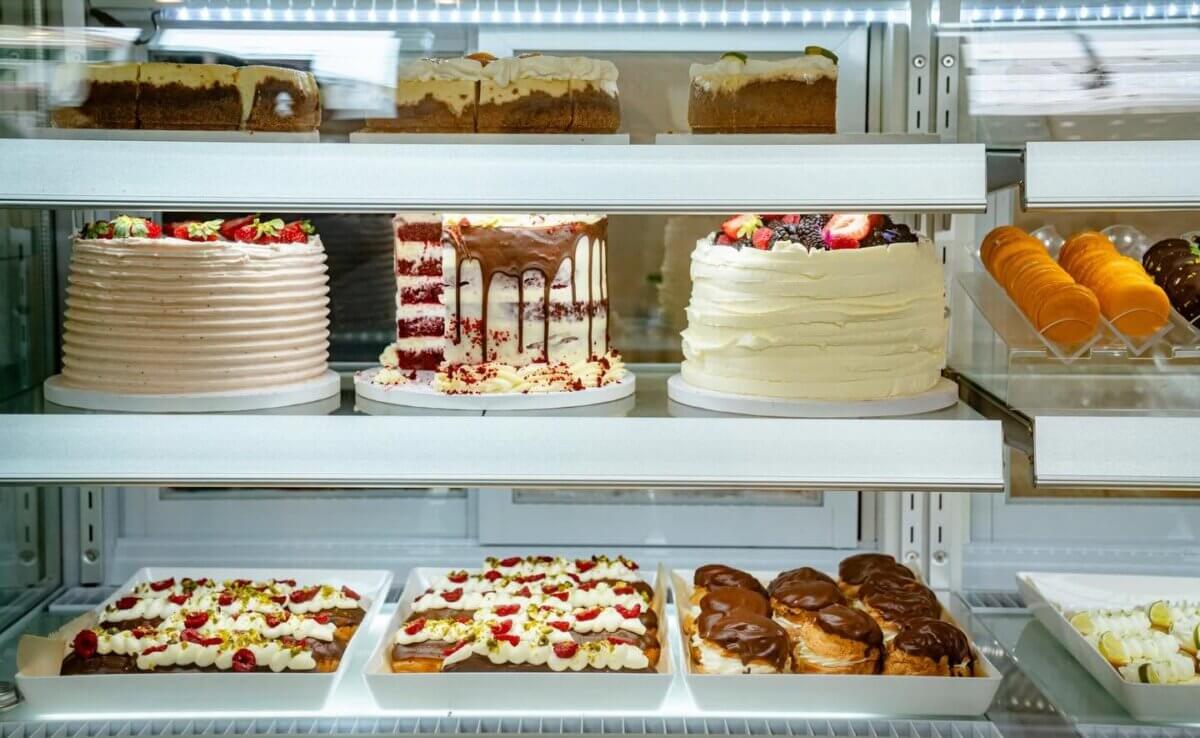 A refrigerated display case with assorted cakes, pastries, and desserts on trays and plates—including layered cakes, eclairs, and fruit-topped treats—organized on glass shelves.