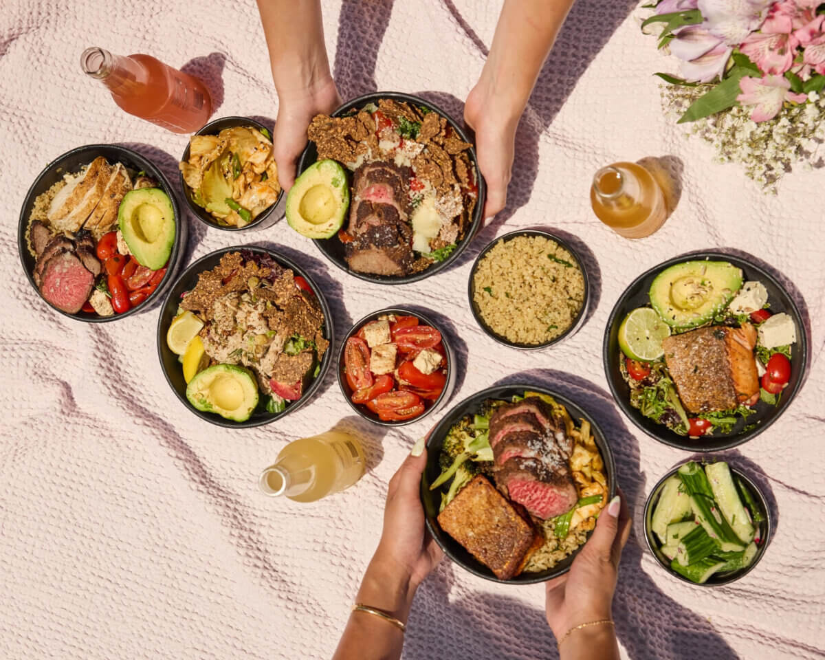 Several hands holding and arranging bowls of salad with steak, avocado, vegetables, and drinks on a pink picnic blanket with flowers in the corner.
