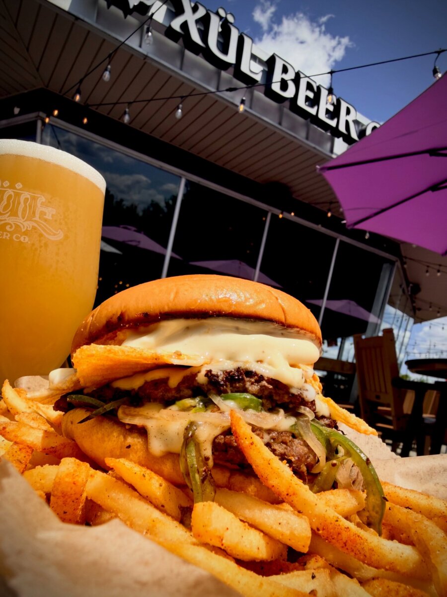 12 New Nashville Restaurants to Try This Month - 18 Cheeseburger topped with fries and melted cheese, served with seasoned French fries and a glass of beer, pictured outside a brewery on a sunny day—a must-try for anyone exploring new Nashville restaurants.