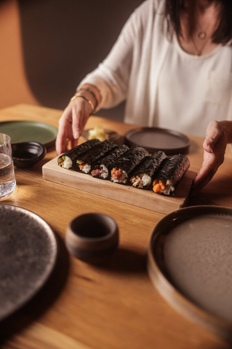 A person places a wooden tray of hand-rolled sushi on a table set with plates, a cup, and a glass of water.