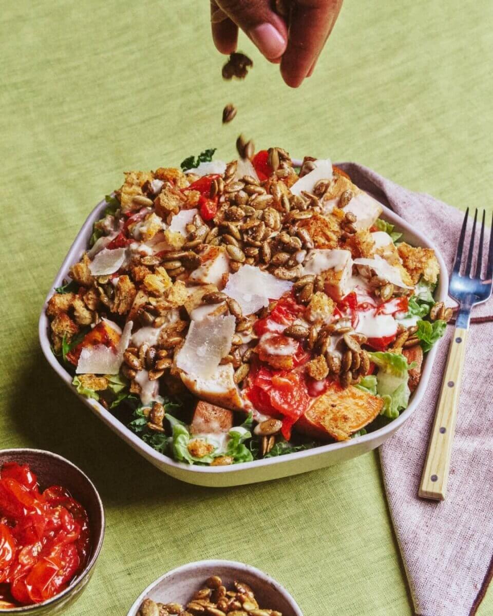 A hand sprinkles seeds over a salad with lettuce, croutons, sliced tomatoes, shaved cheese, and dressing on a green tablecloth with a fork and napkin beside the bowl—an inviting scene reminiscent of the fresh flavors debuting at new Nashville restaurants in July 2026.