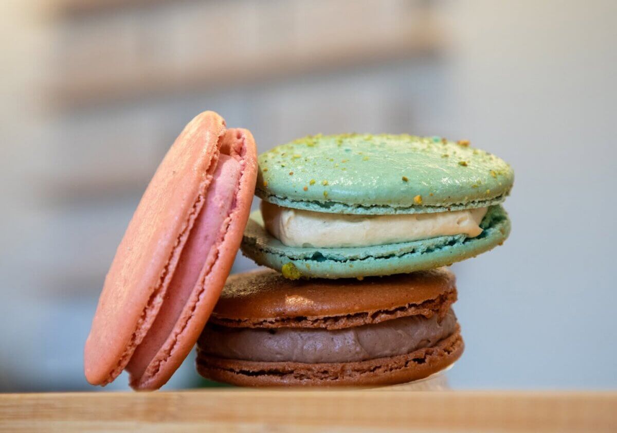 Three macarons—pink, green, and brown with gelato filling—stacked on a wooden surface.