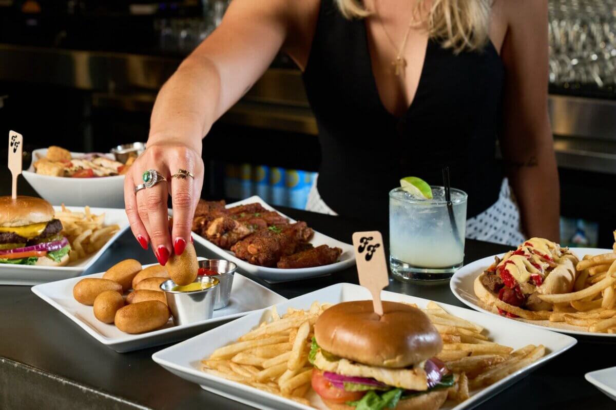 A person reaches for a fried appetizer on a table with burgers, fries, fried chicken, and a cocktail—capturing the lively spirit of new Nashville restaurants arriving in July 2026.