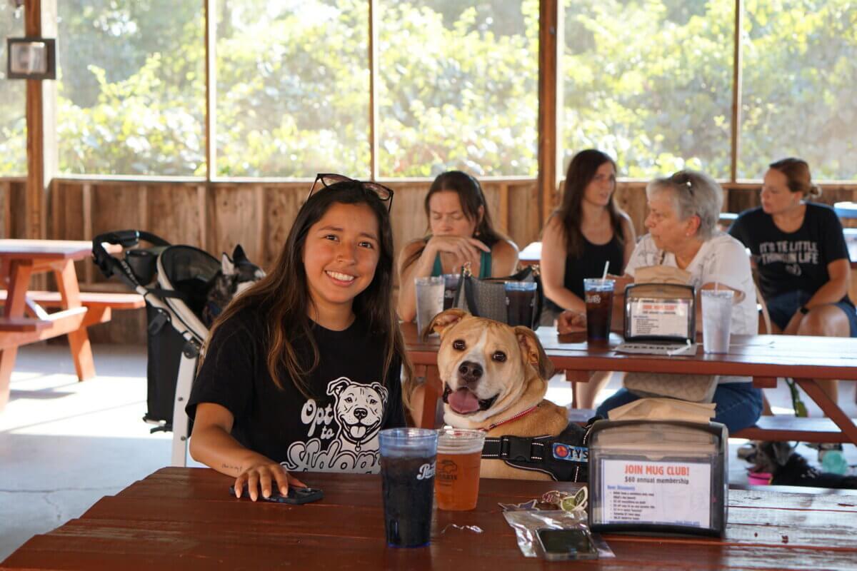 Hey Nashville, Here's Your Chance to Make Your Pet Famous! - 5 A woman and a dog sit at a picnic table in a casual indoor setting with other people in the background, all enjoying drinksβperhaps even raising a toast with an Adoption Ale.