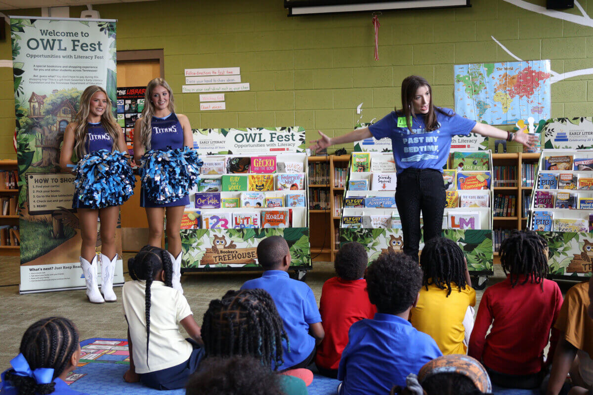 She’s Helping Put Books in the Hands of 500,000 Tennessee Kids - 3 Two cheerleaders and a presenter stand in front of children in a library, with bookshelves and event banners in the background.