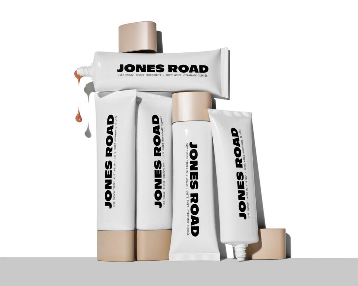 Bobbi Brown's Essential Makeup Musts for Every Decade - 6 Five white Jones Road cosmetic tubes, created by Bobbi Brown, some standing upright and some lying down, are arranged on a flat surface. One tube is open, dispensing a small amount of product.