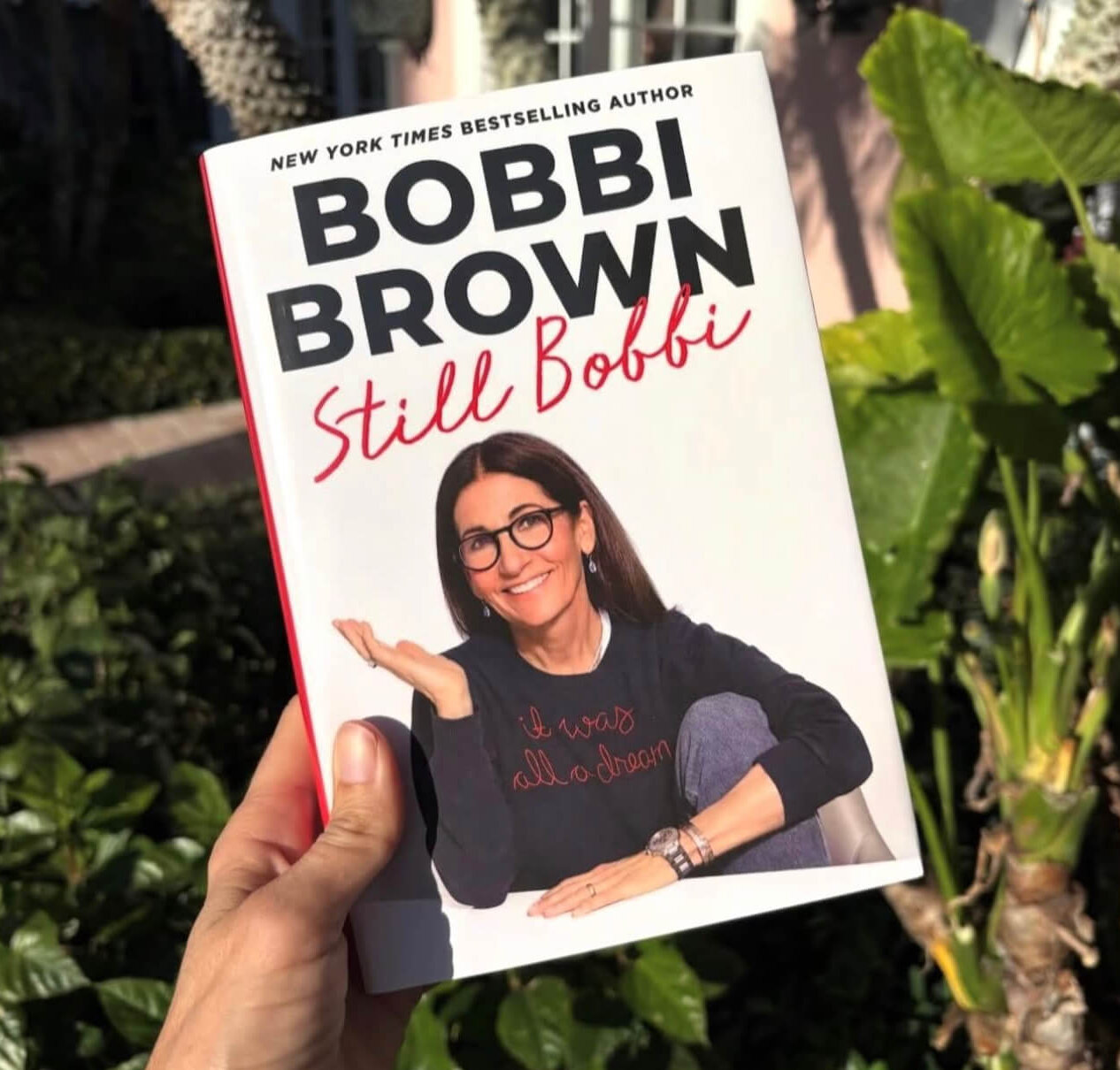 Bobbi Brown's Essential Makeup Musts for Every Decade - 5 A hand holds the book "Still Bobbi" by Bobbi Brown outdoors, with green leaves and sunlight in the background.
