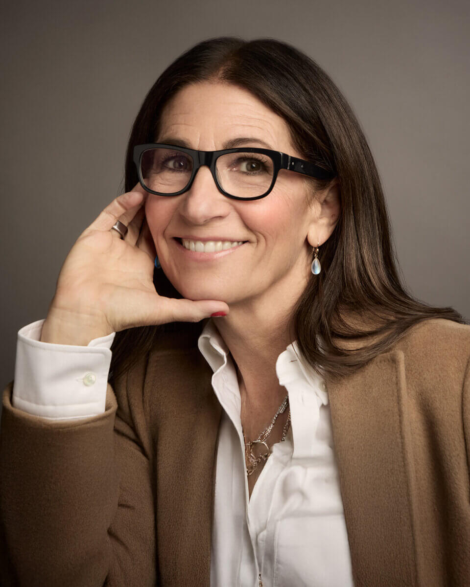Bobbi Brown's Essential Makeup Musts for Every Decade - 2 Woman with long brown hair and glasses, wearing a white shirt and tan blazer, smiling and resting her head on her hand against a neutral backgroundβeffortlessly embodying beauty at every age.