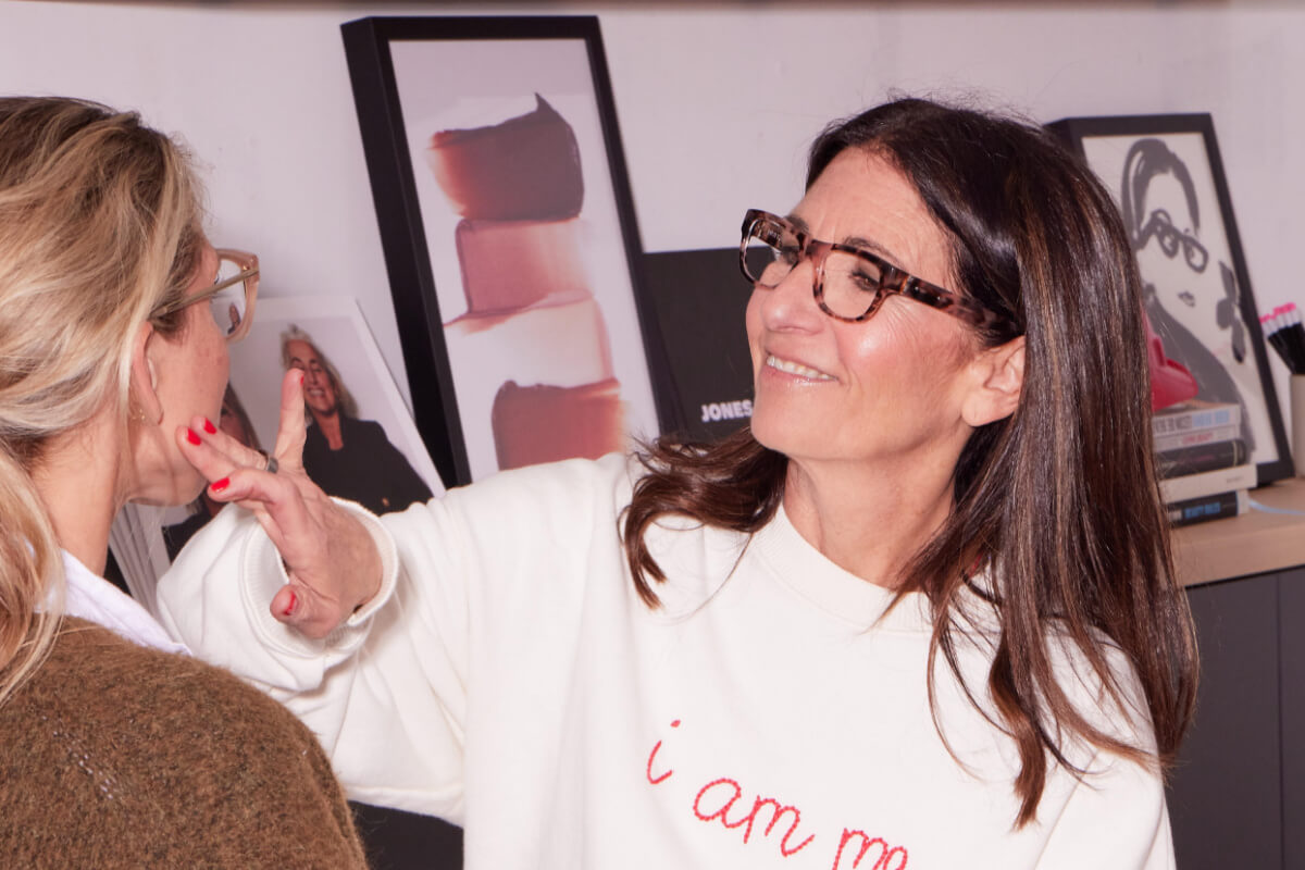 Makeup artist Bobbi Brown wearing glasses and a white shirt applies makeup to a blonde woman facing her.