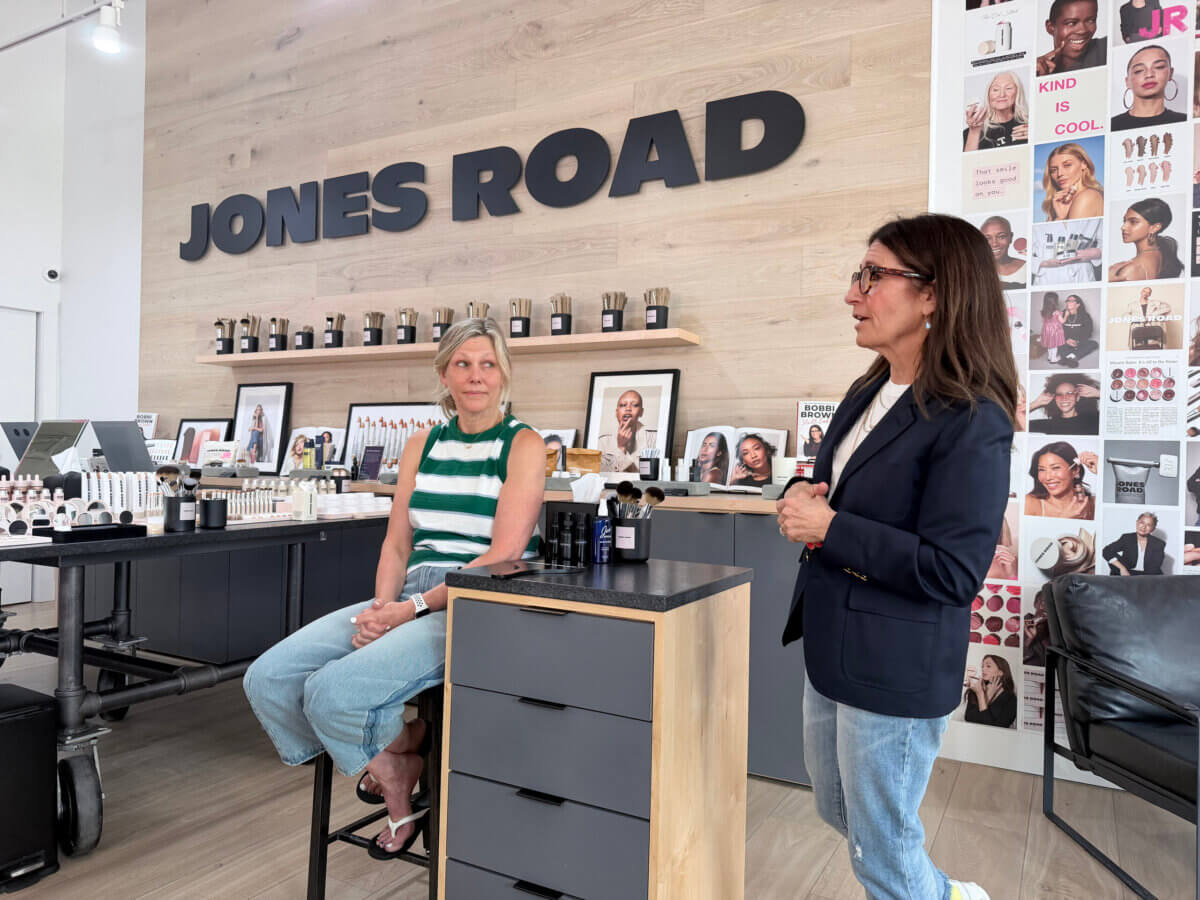 Bobbi Brown's Essential Makeup Musts for Every Decade - 4 Two women are in a makeup store with "JONES ROAD" on the wall; one sits on a stool while the other stands and speaks. Various cosmetics and photos are displayed around them.