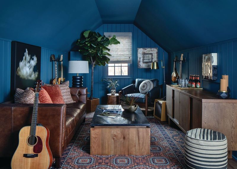 This Designer Wrote the Book on Creating Colorful Spaces — Literally! - 5 A cozy living room with blue walls, leather sofa, striped armchair, wooden furniture, guitars, art, potted plant, patterned rug, and various decor items.