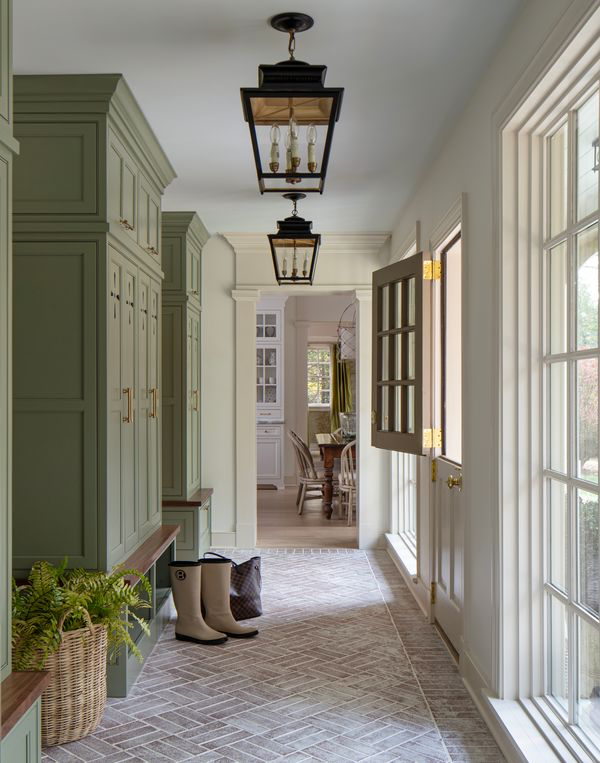 This Designer Wrote the Book on Creating Colorful Spaces — Literally! - 16 A hallway with green cabinets, a wooden bench, wicker basket with fern, rain boots, and a bag on a patterned floor, lantern-style ceiling lights overhead, and an open door leading to the dining room.