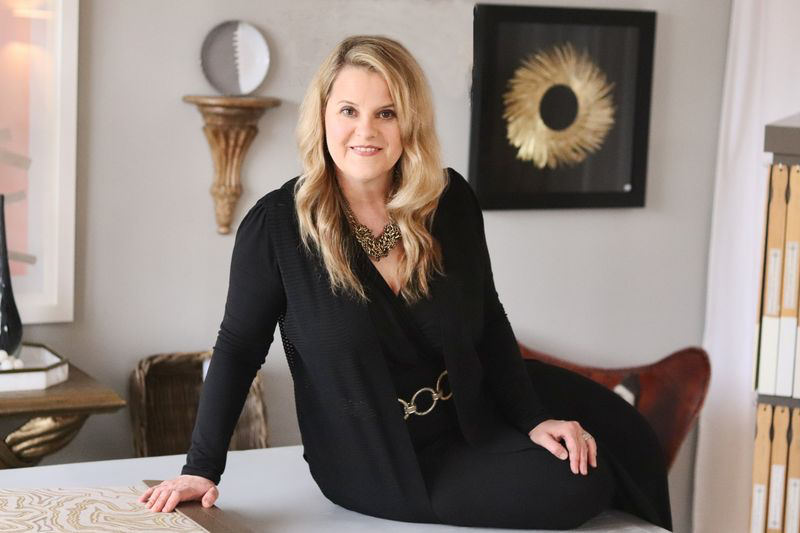 This Designer Wrote the Book on Creating Colorful Spaces — Literally! - 2 A designer with long blonde hair in a black outfit sits on a table in a modern, stylish office filled with decorative art and colorful spaces.