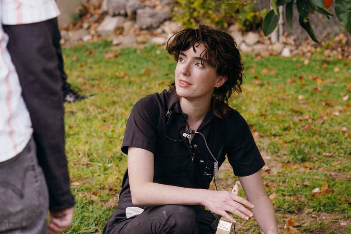 Meet the Memphis Designer Behind the Eerie World of "Obsession" - 4 A Memphian in dark clothing sits on grass, a paintbrush in hand, gazing up at another person standing nearby. Fallen leaves are crafted into a vivid scene around them.