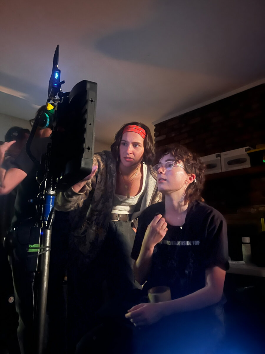 Meet the Memphis Designer Behind the Eerie World of "Obsession" - 3 Two people focus on a monitor—one standing and pointing, the other seated with a cup—in a dimly lit room lined with equipment.