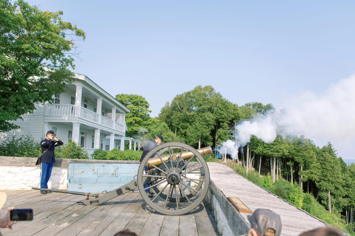 Midwest Weekend Getaway: My Mackinac Island Escape - 9 A person in historic clothing fires a cannon on a wooden platform next to a large white building, with smoke visible in the air and trees in the background.