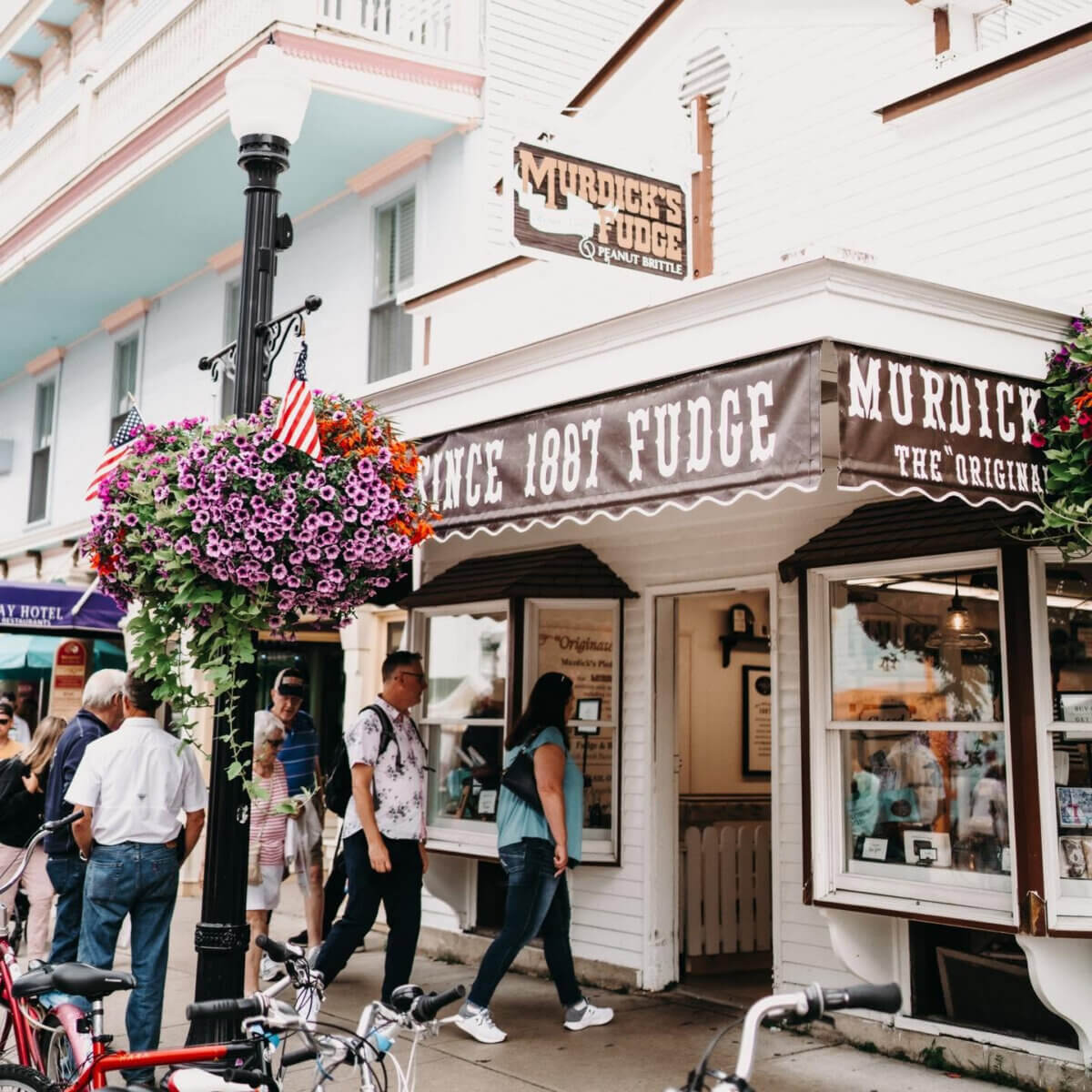 Midwest Weekend Getaway: My Mackinac Island Escape - 10 People walk past Murdick’s Fudge shop, established 1887, with hanging flower baskets and bicycles parked nearby on a lively street.