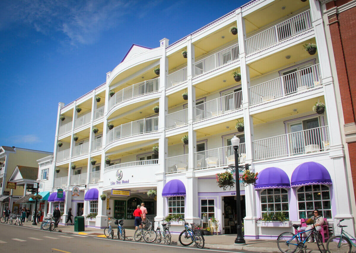Midwest Weekend Getaway: My Mackinac Island Escape - 5 A four-story hotel with white railings, yellow walls, and purple awnings stands on a street with people walking and bikes parked nearby.