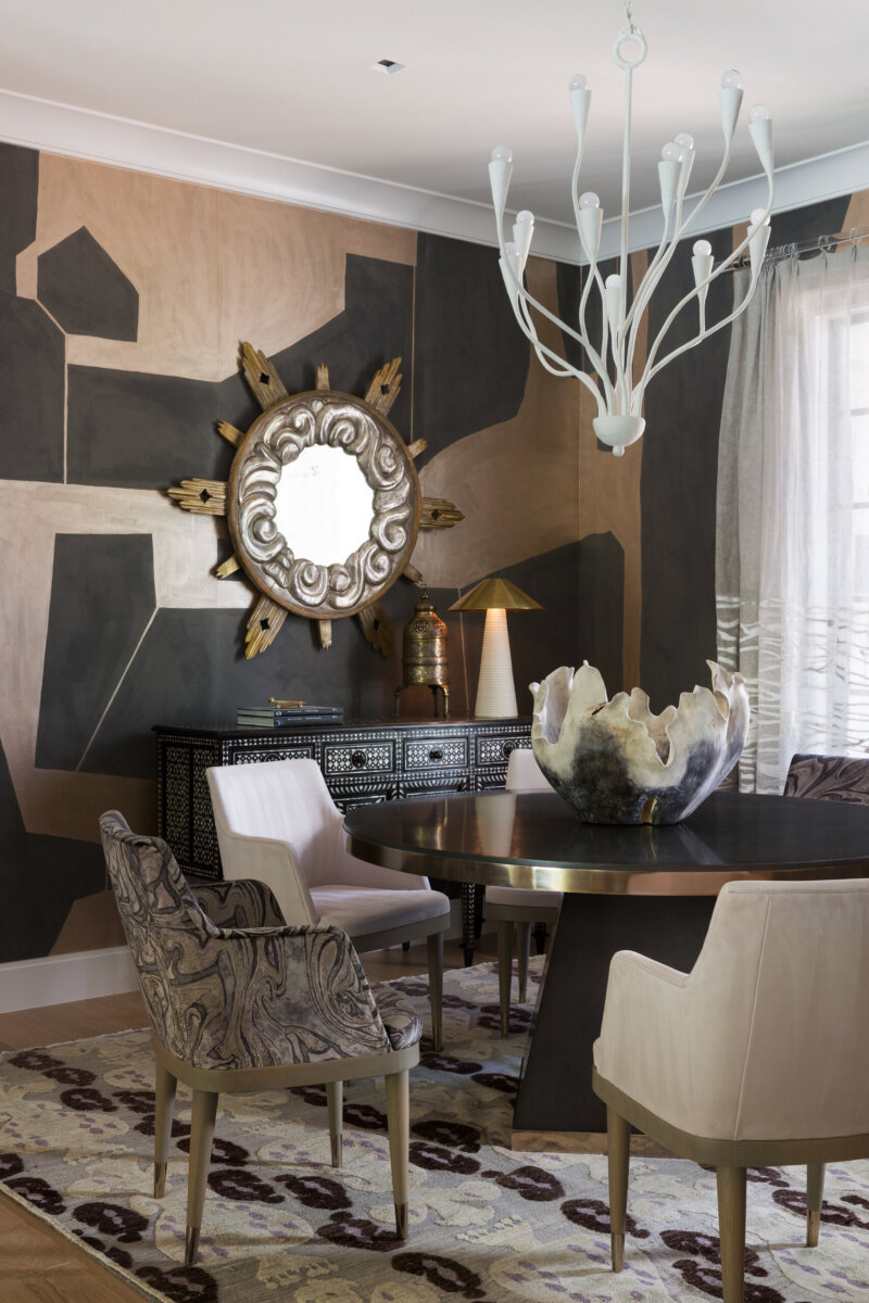 This Designer Wrote the Book on Creating Colorful Spaces — Literally! - 13 A dining room featuring a round table, mixed upholstered chairs, a large decorative bowl, sunburst wall mirror, abstract wall art, chandelier, and patterned rug.