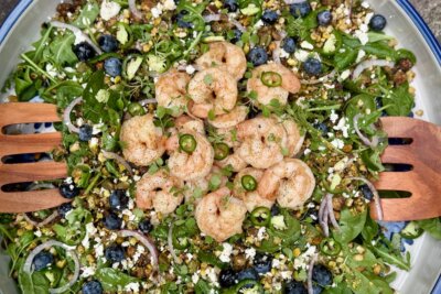A vibrant summer salad, featuring shrimp, fresh greens, blueberries, feta cheese, red onion, sliced jalapeño, avocado, and wholesome grains—all beautifully served with wooden salad tongs.