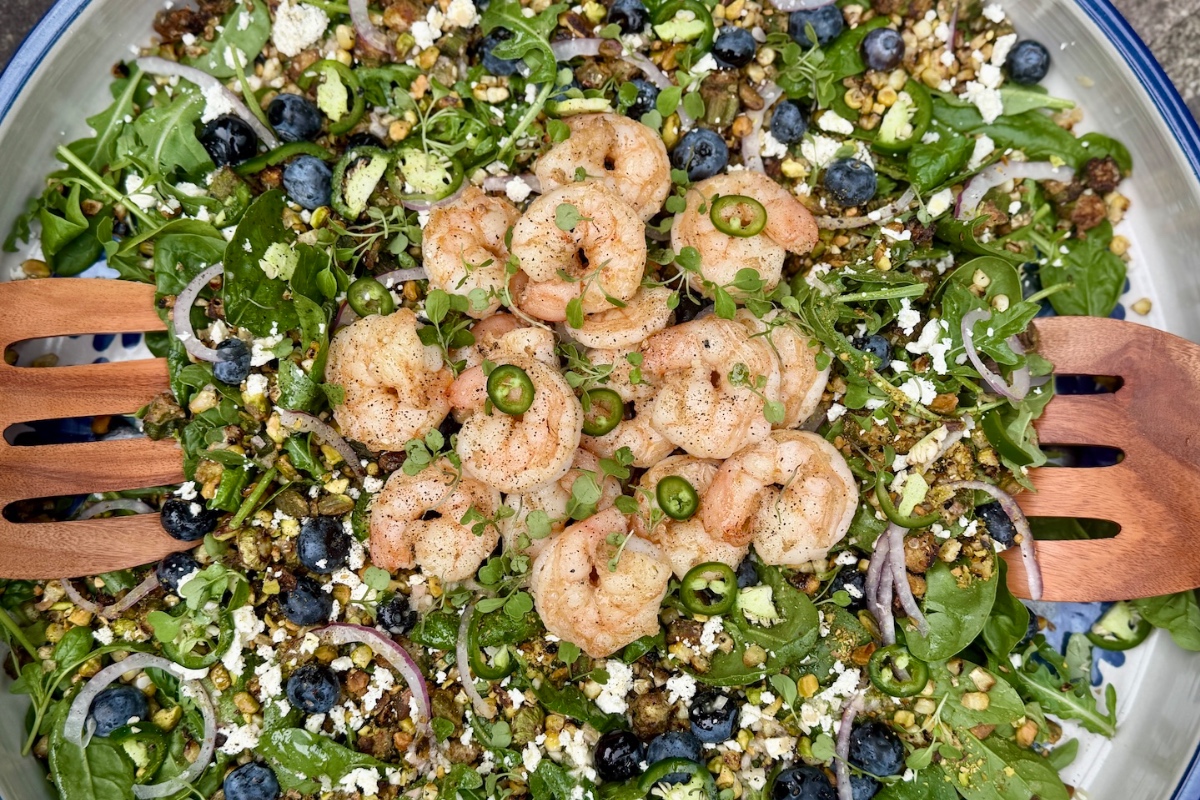 A vibrant summer salad, featuring shrimp, fresh greens, blueberries, feta cheese, red onion, sliced jalapeño, avocado, and wholesome grains—all beautifully served with wooden salad tongs.
