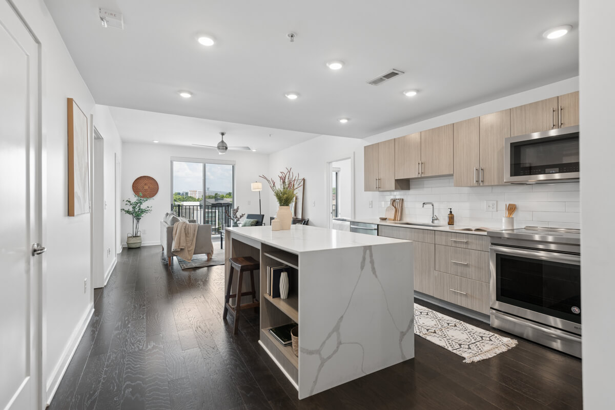 Modern open-concept kitchen and living area at 3116 W End Cir, Nashville TN, featuring a marble island, light wood cabinets, stainless steel appliances, and large windows leading to a balcony.
