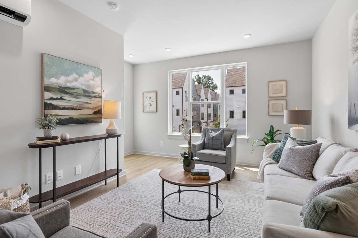 Bright living room with modern decor, a large window, light-colored furniture, a coffee table, wall art, and a view of neighboring buildings outside—perfectly capturing the contemporary charm of East Village Nashville at 323 East Village Ln.