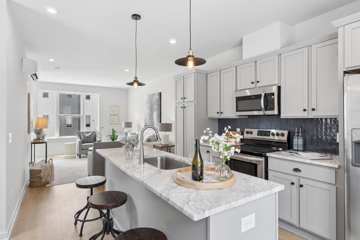 Modern kitchen at 323 East Village Ln with gray cabinets, marble island, stainless steel appliances, pendant lights, and stools. Living area with armchair and window visible in the background—experience stylish comfort in the heart of East Village Nashville.