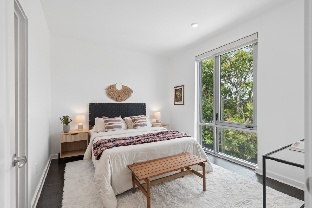 Bright bedroom in Nashville TN with a large window, double bed with striped pillows, two bedside tables with lamps, a wooden bench at the foot of the bed, and a white rug on dark flooring at 3116 W End Cir.