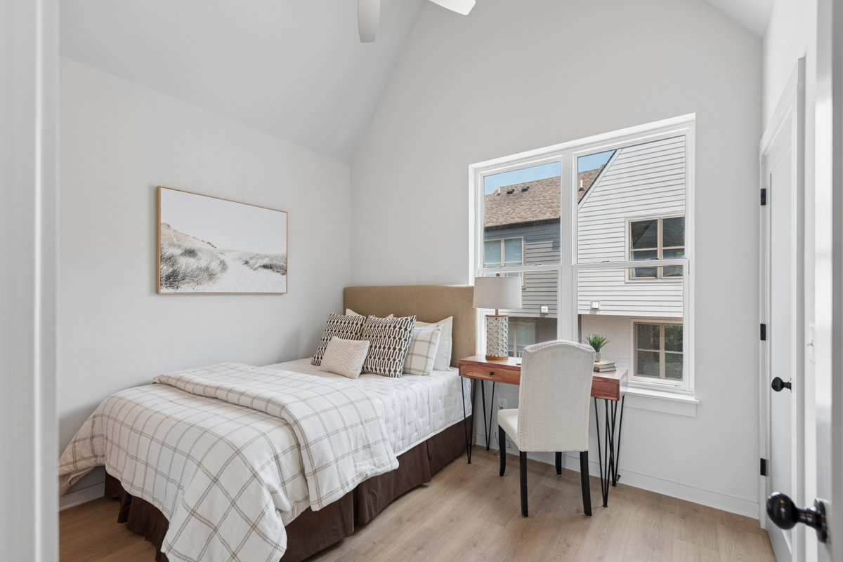 A small bedroom with a neatly made bed, a wooden desk and chair by a window, light walls, and minimal decor offers a cozy retreat within the desirable East Village Nashville community.