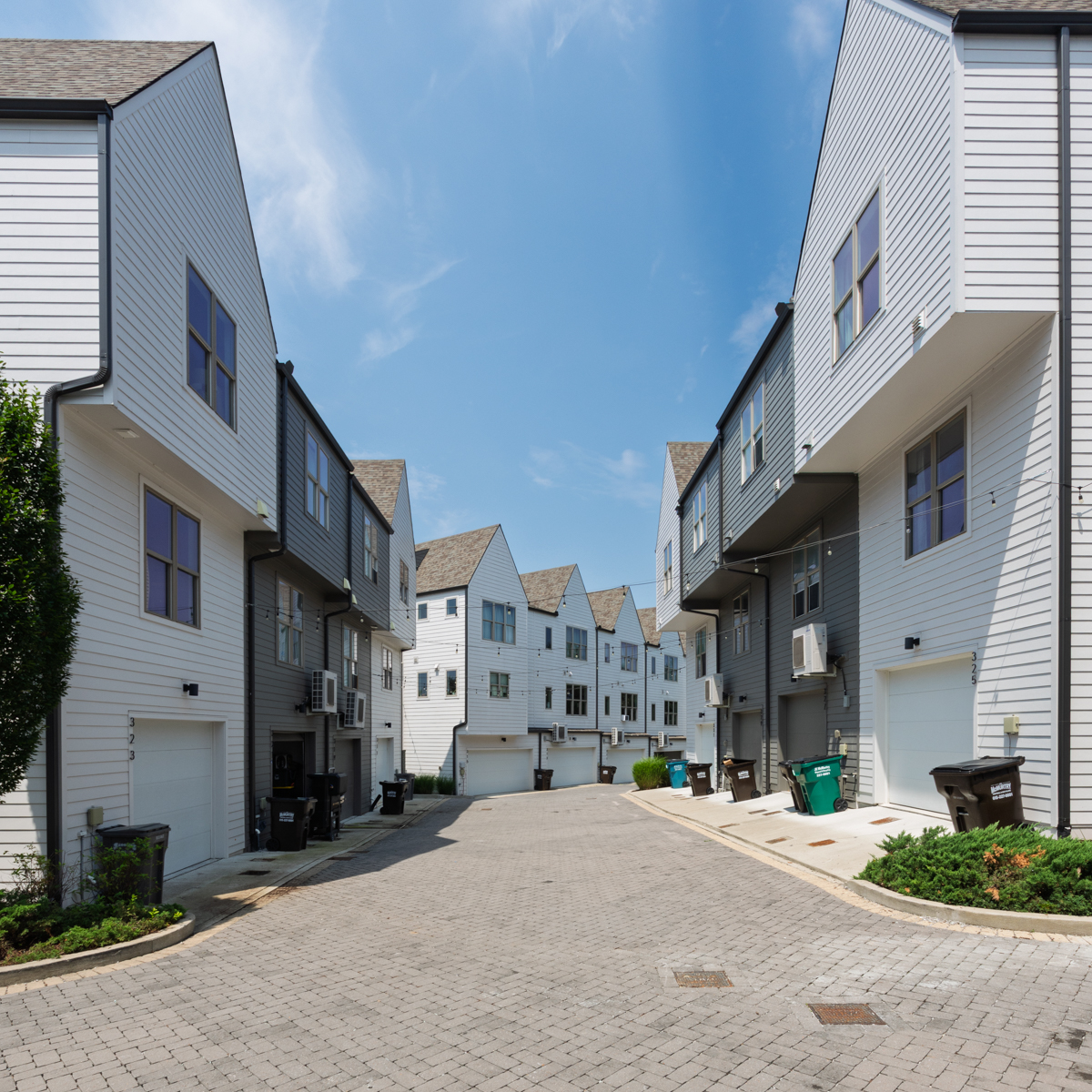 A row of modern townhouses with garages faces each other along a paved alley at 323 East Village Ln, Nashville, under a blue sky with scattered clouds.