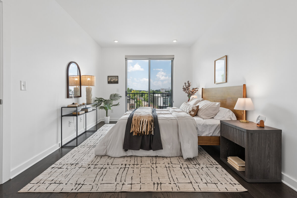 A modern bedroom in Nashville TN featuring a wooden bed, bedside table, console table, and large window overlooking a balcony with a city view. Neutral tones and minimal decor at 3116 W End Cir create a clean, inviting look.