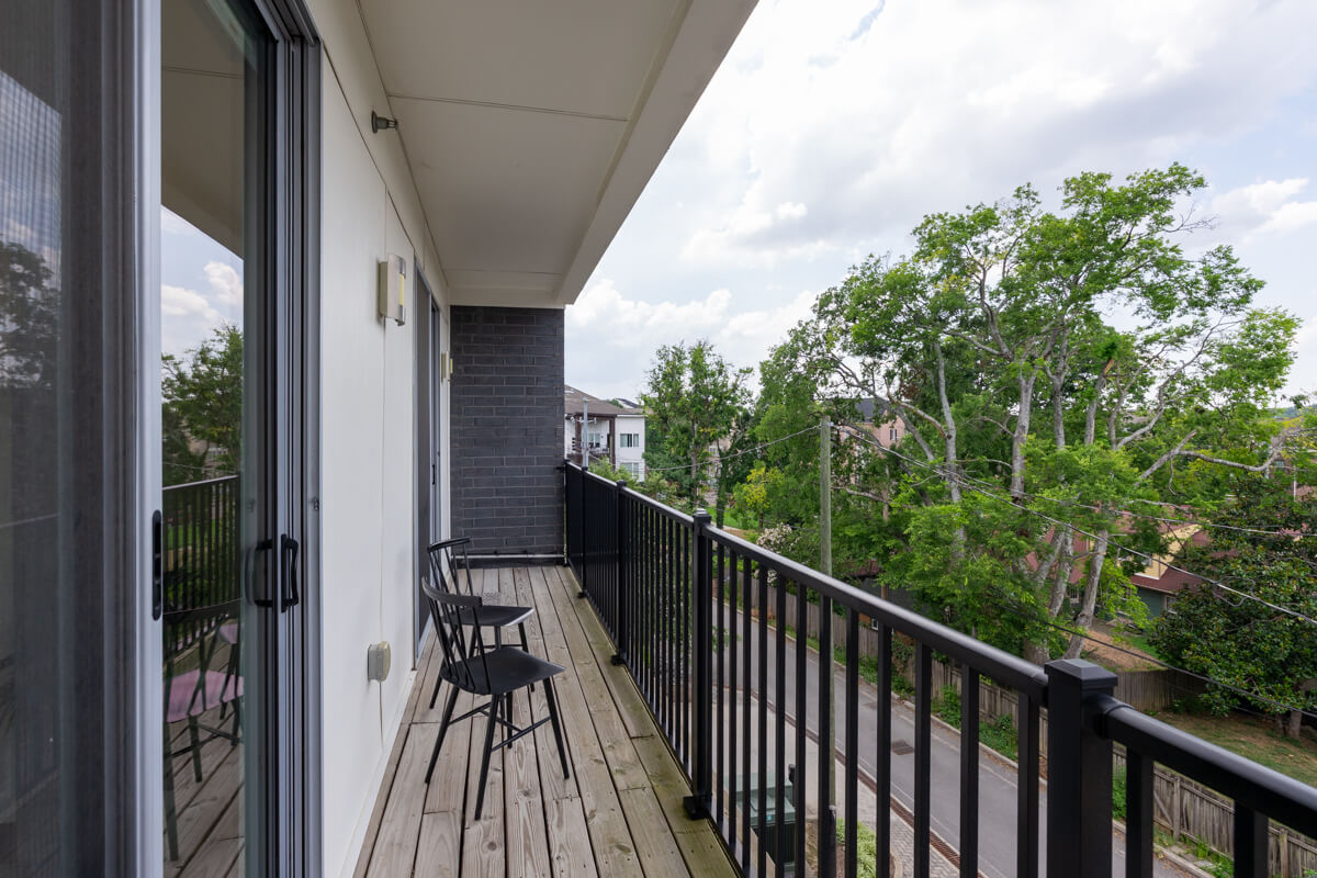 A modern apartment balcony at 3116 W End Cir, Nashville TN, features two black chairs, a small table, wooden flooring, and a black railing, overlooking a street and green trees.