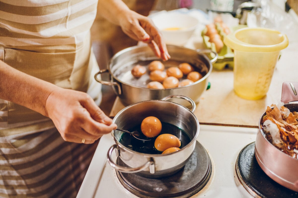 Person in a striped apron boils eggs in two pots on a stove, using a spoon to handle eggs—preparing complete protein foods to help meet daily protein goals, with an egg carton and kitchen items nearby.