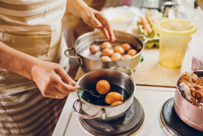 Person in a striped apron boils eggs in two pots on a stove, using a spoon to handle eggs—preparing complete protein foods to help meet daily protein goals, with an egg carton and kitchen items nearby.